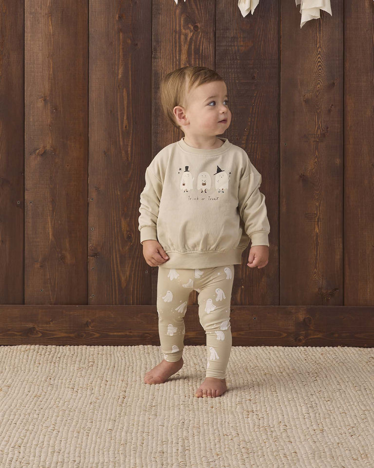 A toddler stands on a textured beige rug in front of a wooden wall, wearing the quincy mae relaxed fleece sweatshirt in trick or treat—crafted from organic cotton with playful animal faces—and matching leggings with white animal patterns, looking up to the side.