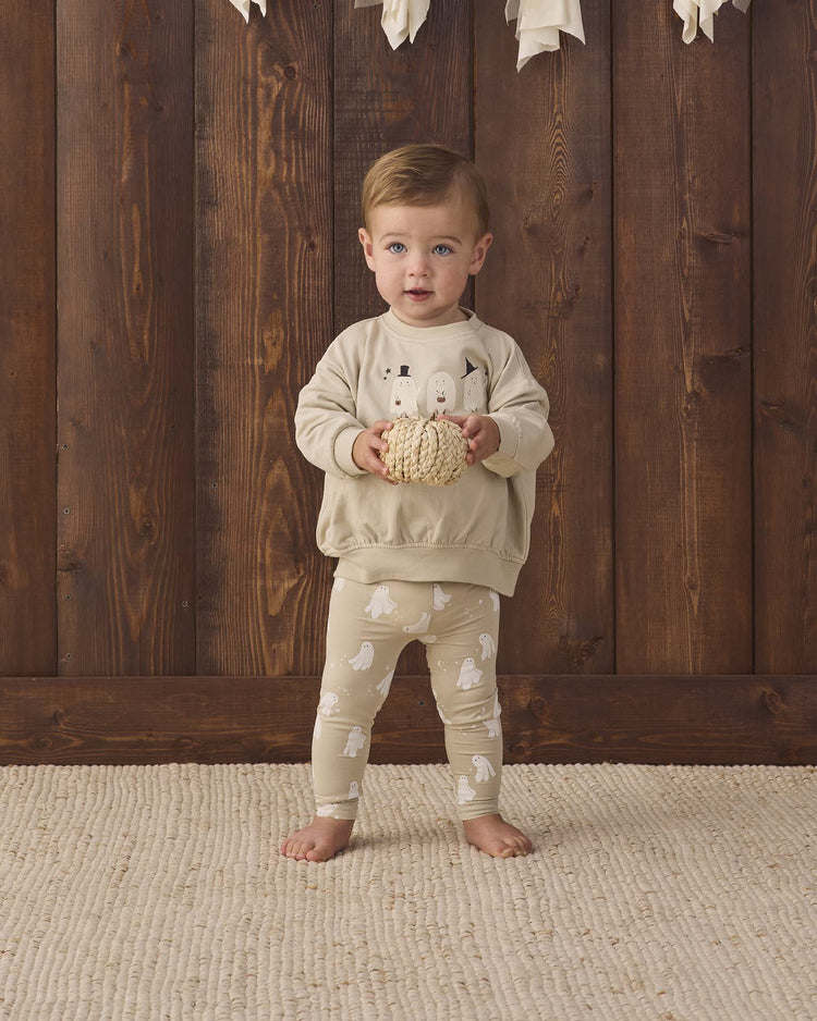 A toddler stands barefoot on a cream rug, wearing the quincy mae relaxed fleece sweatshirt in trick or treat, featuring playful trick or treat print and white ghost designs, holding a textured ball in front of a wooden wall with white decorations hanging above.