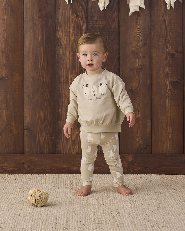 A toddler stands on a textured rug in front of a wooden wall, wearing the quincy mae relaxed fleece sweatshirt in trick or treat. A small decorative ball is on the floor nearby.