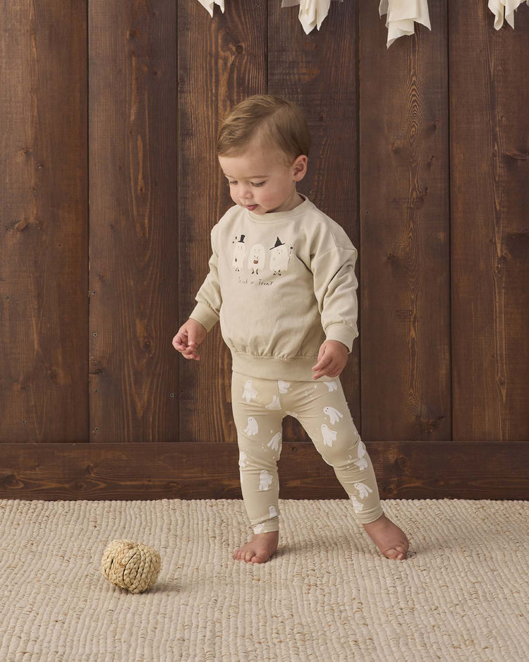 A young child wearing the quincy mae relaxed fleece sweatshirt in trick or treat stands on a cream rug next to a textured ball, with a wooden wall in the background.