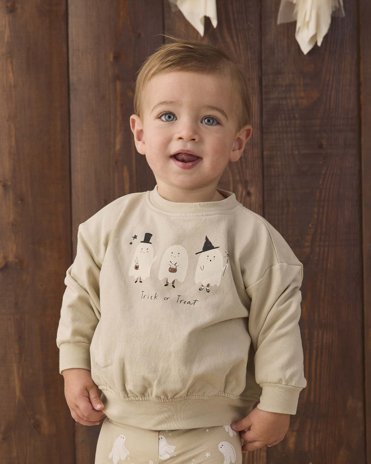 A toddler with light brown hair wears the quincy mae relaxed fleece sweatshirt in trick or treat, made from organic cotton and featuring playful ghosts and a fun trick or treat print, standing in front of a wooden background.