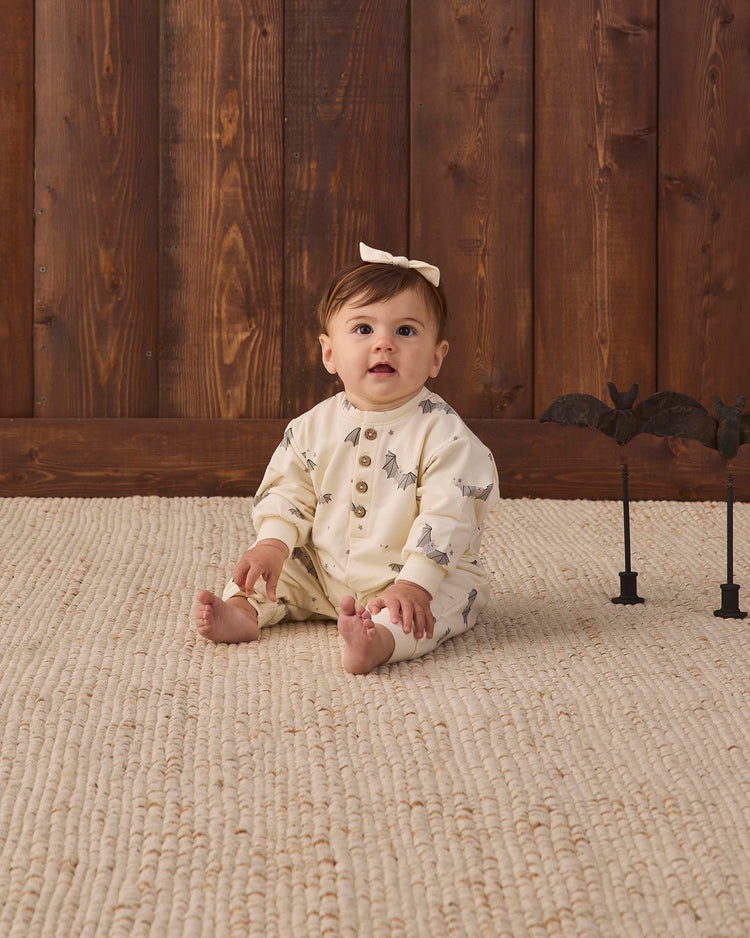 A baby in a quincy mae relaxed fleece jumpsuit in baby bats sits on a textured rug in front of a wooden wall, wearing a bow headband. Two black bat decorations stand to the right.
