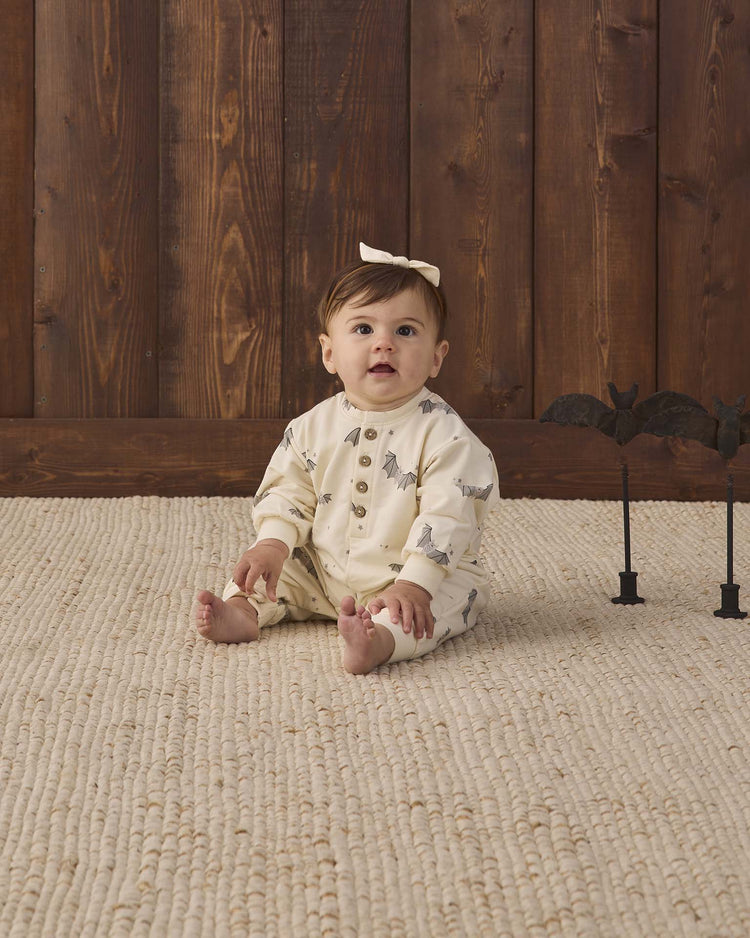 A baby in a quincy mae relaxed fleece jumpsuit in baby bats sits on a textured rug in front of a wooden wall, wearing a bow headband. Two black bat decorations stand to the right.