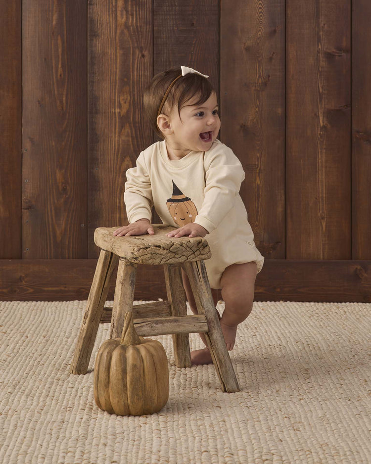 A smiling baby wearing the quincy mae crewneck bubble romper in lil pumpkin stands holding a rustic wooden stool, with a small pumpkin on the floor. The background features wood paneling and a woven rug.