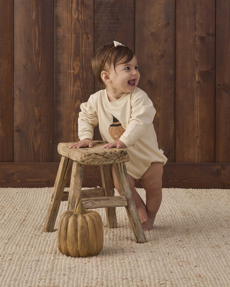 A smiling baby stands holding a rustic wooden stool, dressed in the quincy mae crewneck bubble romper in lil pumpkin. A decorative pumpkin sits on the floor in front, with wooden paneling in the background.
