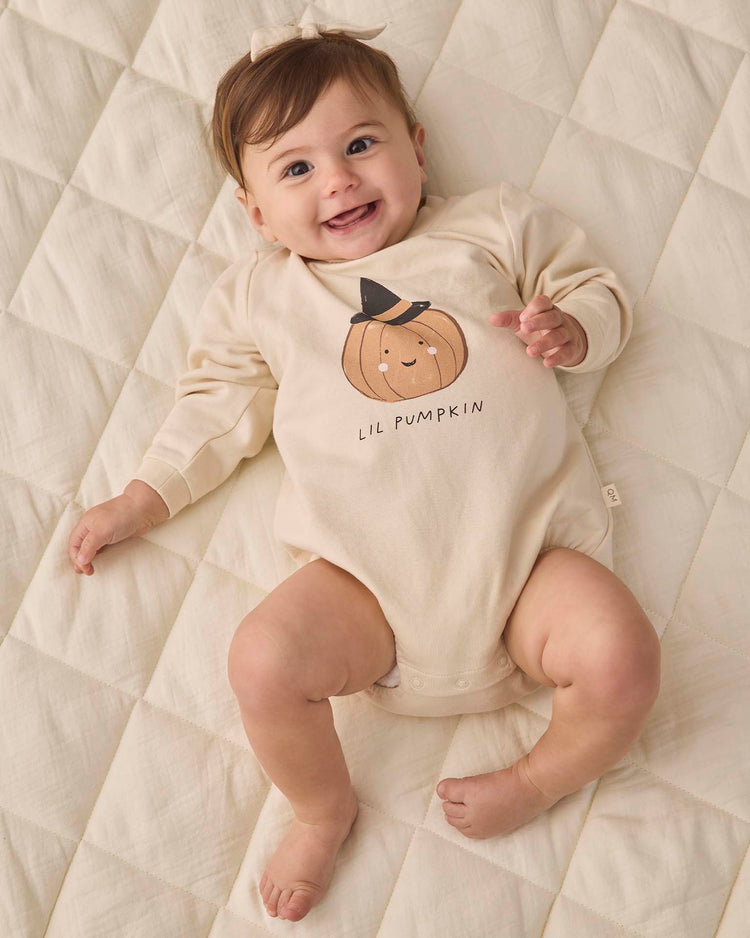 A smiling baby lies on a quilted cream blanket, wearing the quincy mae crewneck bubble romper in lil pumpkin.