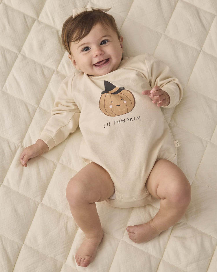 A smiling baby lies on a quilted cream blanket, wearing the quincy mae crewneck bubble romper in lil pumpkin.