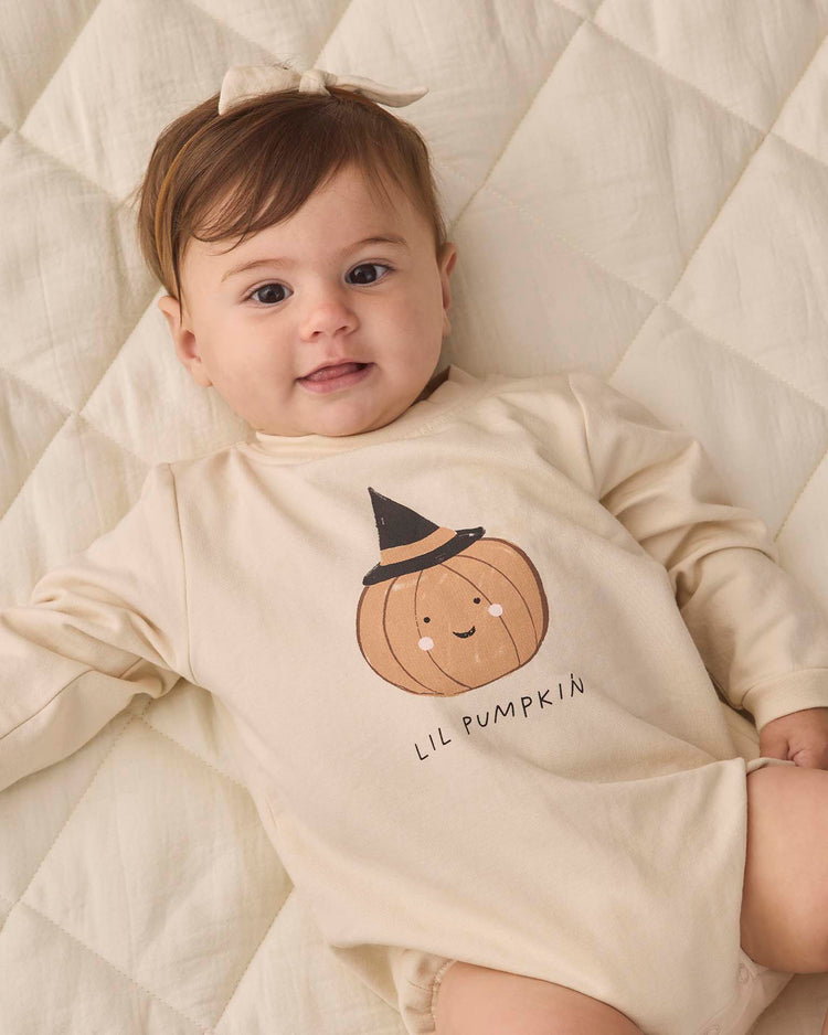 A baby lies on a quilted cream blanket, dressed in the quincy mae crewneck bubble romper in lil pumpkin—an organic cotton beige romper featuring a pumpkin and witch hat graphic labeled “Lil Pumpkin”—paired with a matching bow headband.