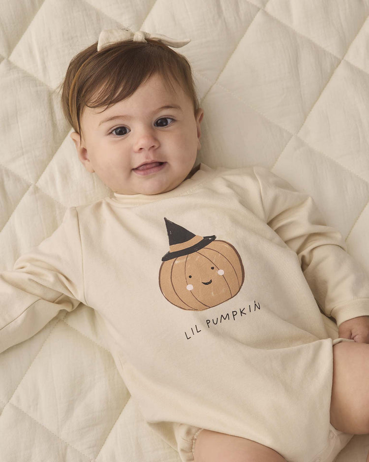 A baby lies on a quilted cream blanket, dressed in the quincy mae crewneck bubble romper in lil pumpkin—an organic cotton beige romper featuring a pumpkin and witch hat graphic labeled “Lil Pumpkin”—paired with a matching bow headband.