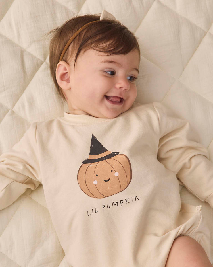 A smiling baby lies on a quilted cream blanket, wearing the quincy mae crewneck bubble romper in lil pumpkin—an organic cotton romper in beige featuring a pumpkin graphic with a cute face and witch hat, and “LIL PUMPKIN” printed below.