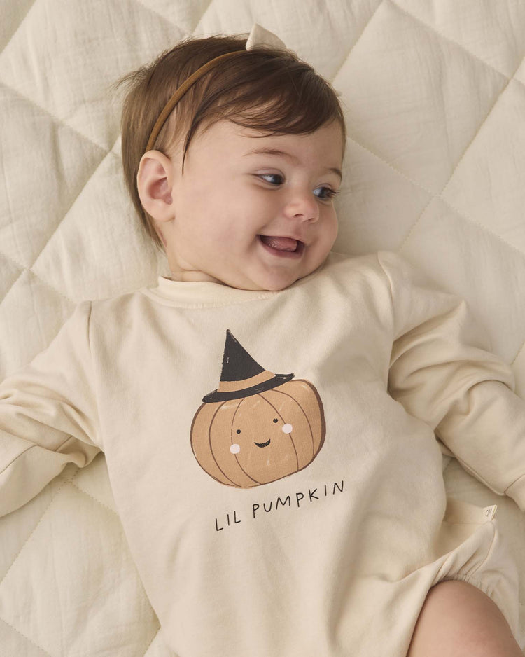 A smiling baby lies on a quilted cream blanket, wearing the quincy mae crewneck bubble romper in lil pumpkin—an organic cotton romper in beige featuring a pumpkin graphic with a cute face and witch hat, and “LIL PUMPKIN” printed below.