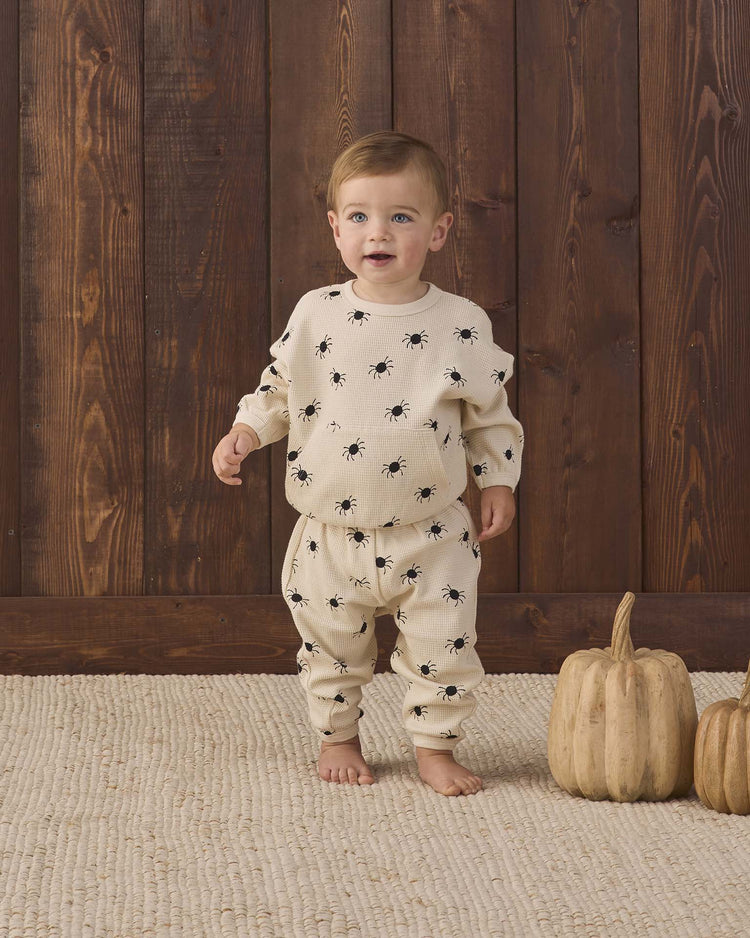 A toddler stands barefoot on a textured rug, wearing the quincy mae waffle slouch set in bitsy spider—a cream organic cotton ensemble decorated with black spider prints. Two light-colored pumpkins rest nearby against a wooden wall.