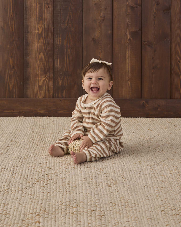 A smiling baby in the Quincy Mae waffle long sleeve jumpsuit in spice stripe, featuring adorable coconut buttons and a sweet bow, sits on a textured beige rug holding a small decorative ball, with a wooden wall in the background.