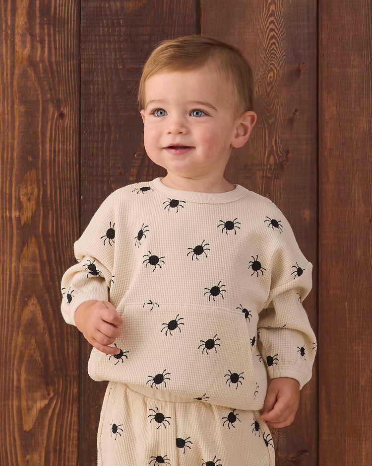 A toddler with light brown hair wears the quincy mae waffle slouch set in bitsy spider—a cream-colored kids loungewear set adorned with black bug prints—while standing in front of a wooden wall, smiling slightly and looking off to the side.