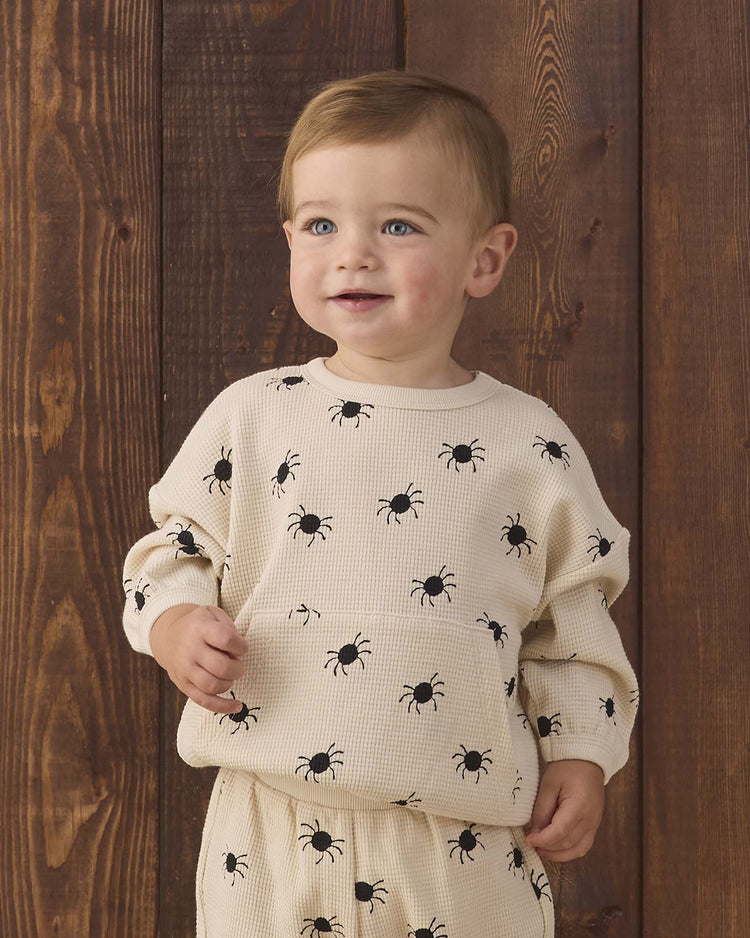 A toddler with light brown hair wears the quincy mae waffle slouch set in bitsy spider—a cream-colored kids loungewear set adorned with black bug prints—while standing in front of a wooden wall, smiling slightly and looking off to the side.