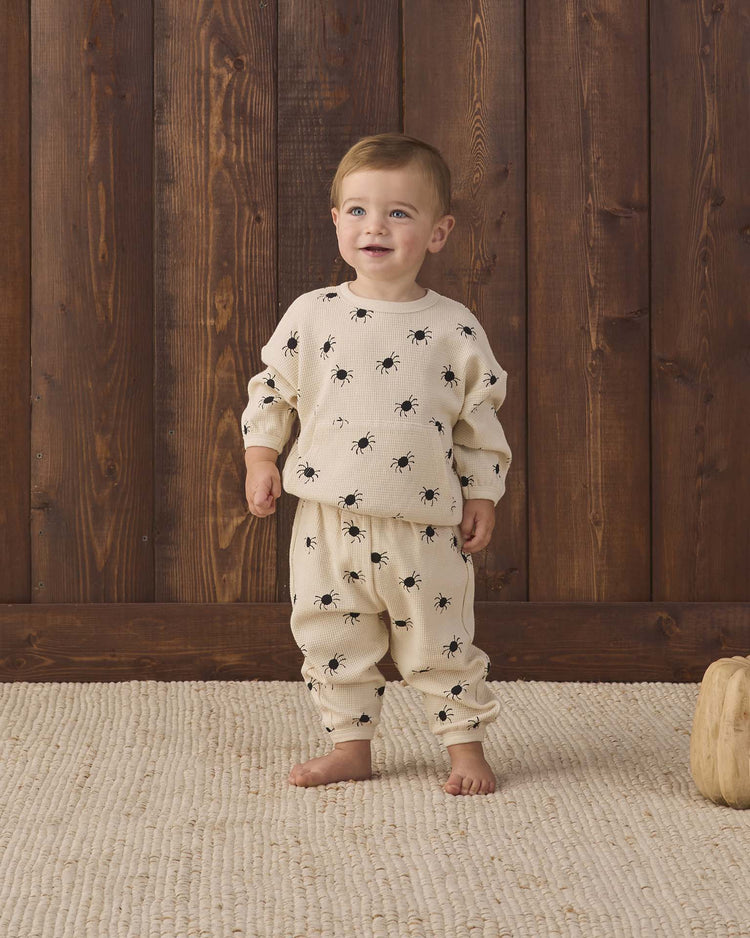 A toddler stands barefoot on a textured rug, wearing the quincy mae waffle slouch set in bitsy spider—a cream-colored kids loungewear set featuring a black spider print, crafted from soft organic cotton—in front of a wooden wall.