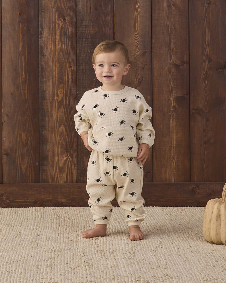A toddler stands on a beige rug, wearing Quincy Mae’s waffle slouch set in bitsy spider—a cream-colored kids loungewear set adorned with playful black bug prints. Crafted from soft organic cotton, this outfit ensures maximum playtime comfort. Wooden panels form the background, with two small wooden pumpkins placed nearby on the floor.