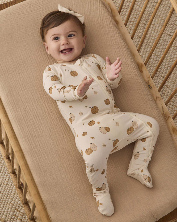 A smiling baby wearing the quincy mae bamboo zip footie in harvest friends—a light-colored outfit with a brown animal print—and a bow headband, lying on a beige mattress in a wooden crib.