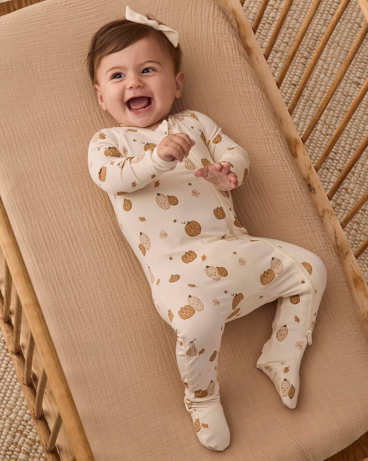 A smiling baby wearing the quincy mae bamboo zip footie in harvest friends lies on a beige crib mattress, reaching forward, with a matching bow on their head.