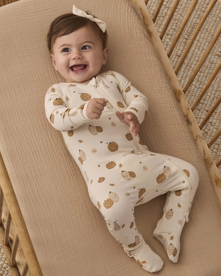 A smiling baby wearing the quincy mae bamboo zip footie in harvest friends, featuring pumpkin and gourd prints, lies on a beige blanket in a wooden crib and wears a matching bow headband.