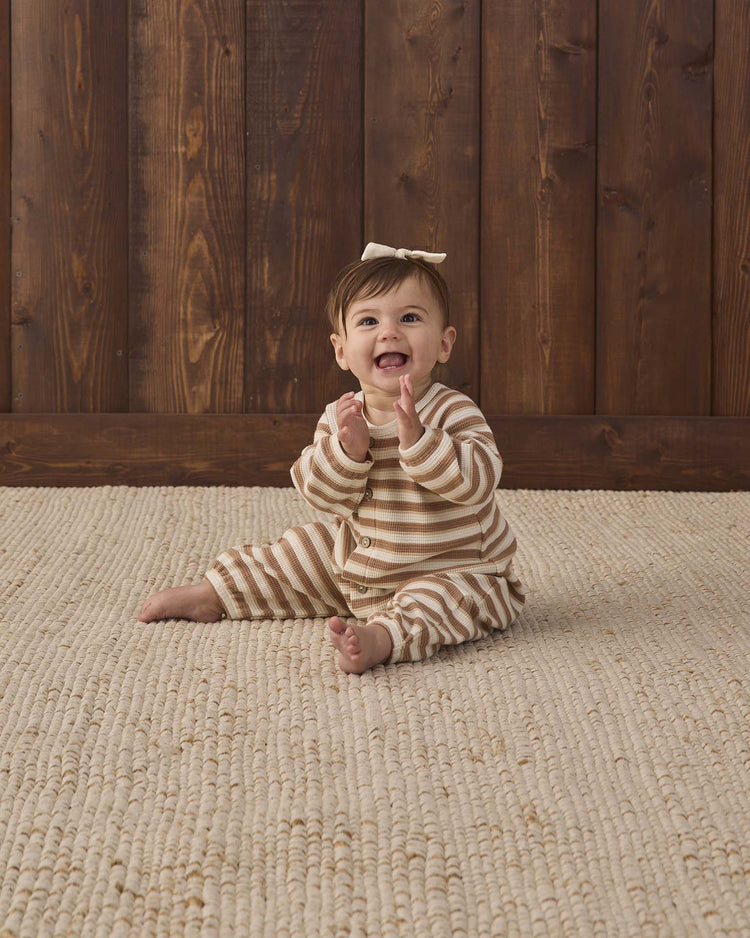 A smiling baby wearing the quincy mae waffle long sleeve jumpsuit in spice stripe, featuring coconut buttons and a bow on their head, sits clapping hands on a textured beige rug in front of a wooden wall.