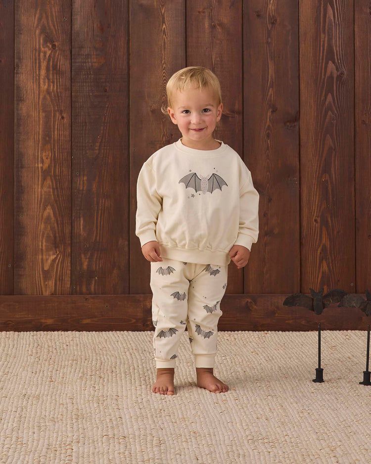 A young child wearing the quincy mae relaxed fleece sweatpant in baby bats stands on a textured rug in front of a wooden wall, smiling at the camera. A black bat decoration is visible on the right side of the image.