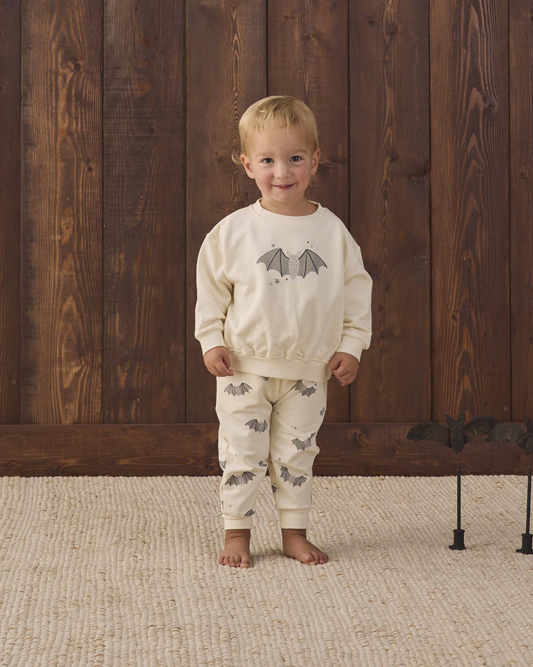 A young child wearing the quincy mae relaxed fleece sweatpant in baby bats stands on a textured rug in front of a wooden wall, smiling at the camera. A black bat decoration is visible on the right side of the image.