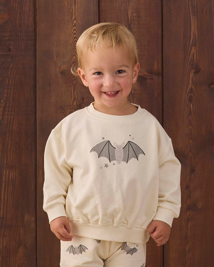 A smiling toddler stands in front of a wooden wall, wearing the quincy mae relaxed fleece sweatshirt in baby bats—a cream-colored organic cotton top adorned with a playful baby bat print—paired with matching pants.
