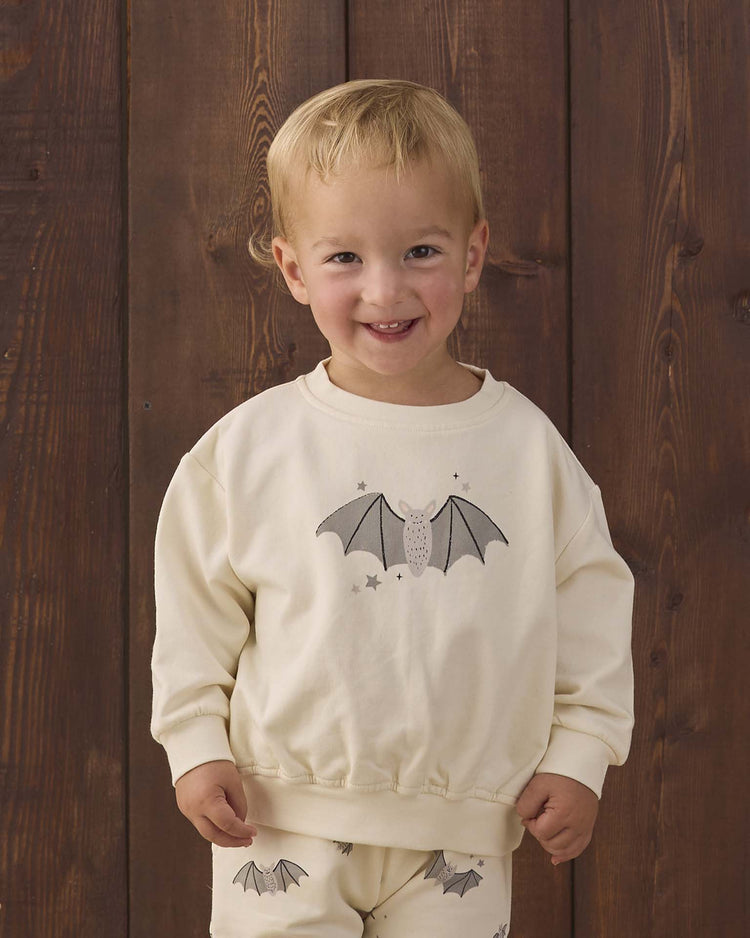 A smiling toddler stands in front of a wooden wall, wearing the quincy mae relaxed fleece sweatshirt in baby bats—a cream-colored organic cotton top adorned with a playful baby bat print—paired with matching pants.