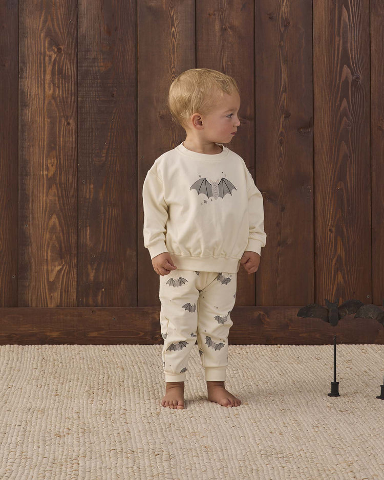 A young child stands on a textured rug in front of a wooden wall, wearing the quincy mae relaxed fleece sweatshirt in baby bats, featuring cream-colored fabric with bat motifs. A toy bat on a stand sits on the floor beside them.