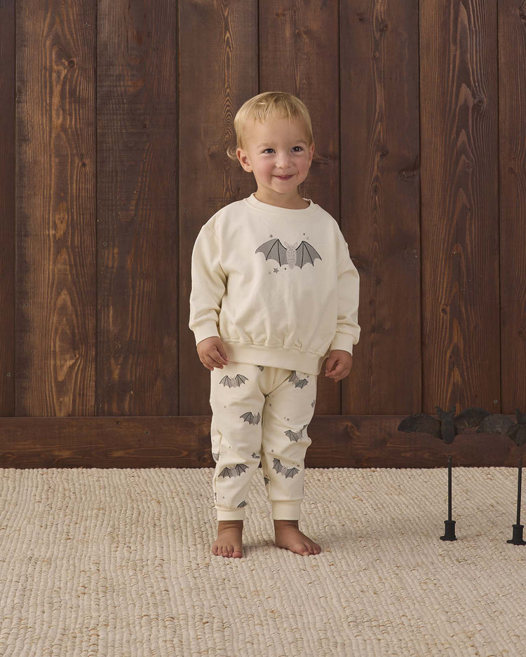 A young child smiles, standing barefoot on a cream rug, wearing the quincy mae relaxed fleece sweatshirt in baby bats—a cozy organic cotton top featuring playful bat prints. Behind them is a wooden wall, with a small bat decoration visible to the right.
