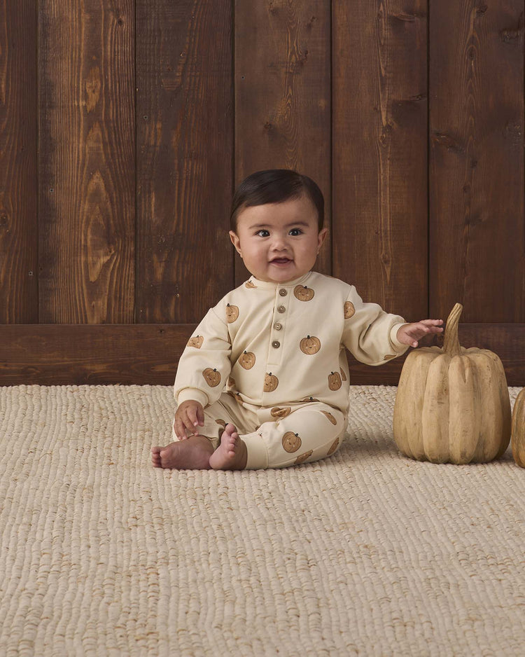 A baby wearing the quincy mae relaxed fleece jumpsuit in pumpkins sits on a textured rug beside a beige pumpkin, with a wooden wall in the background.