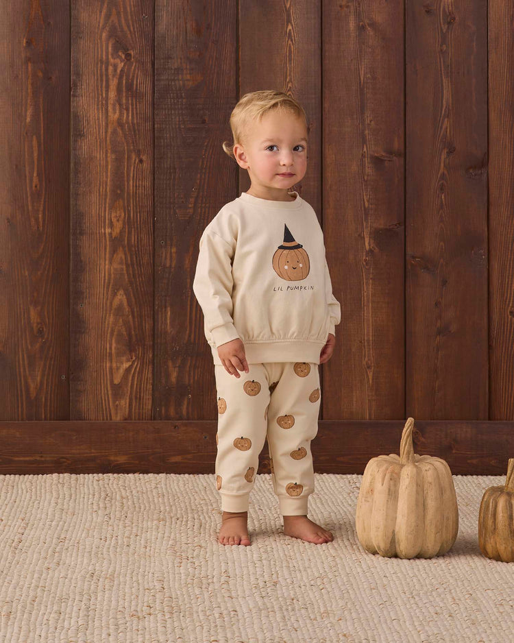 A toddler stands on a cream rug wearing the quincy mae relaxed fleece sweatshirt in lil pumpkin, with two beige pumpkins nearby and a wooden wall in the background, radiating cozy essential vibes perfect for autumn nights.