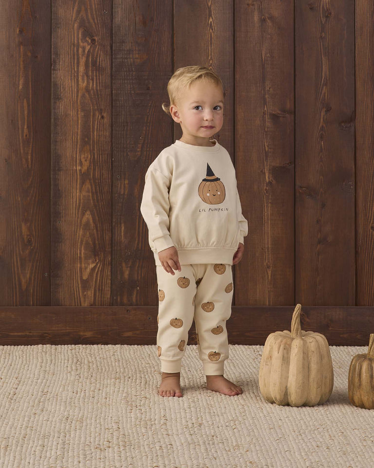 A toddler stands on a cream rug wearing the quincy mae relaxed fleece sweatshirt in lil pumpkin, with two beige pumpkins nearby and a wooden wall in the background, radiating cozy essential vibes perfect for autumn nights.