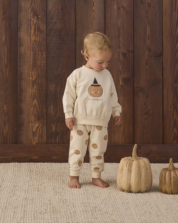 A toddler stands on a cream rug, wearing the quincy mae relaxed fleece sweatshirt in lil pumpkin. Two decorative pumpkins sit beside the child, while a wooden wall in the background adds a warm, rustic feel.