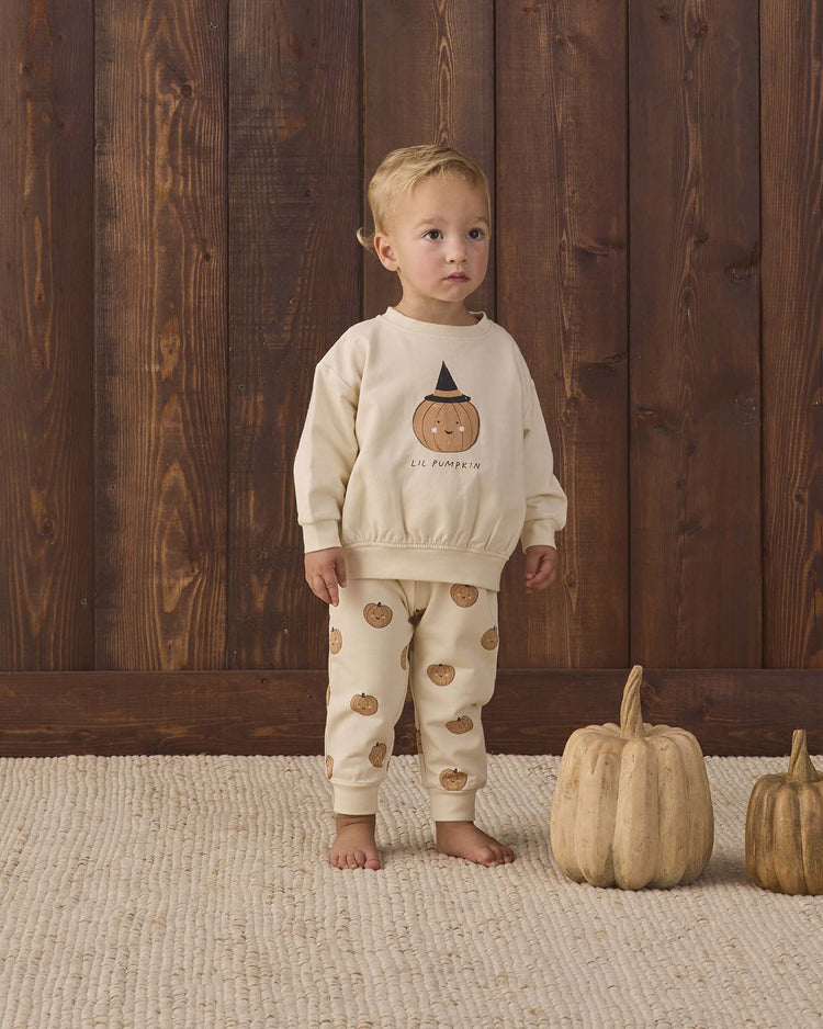 A toddler wearing the Quincy Mae relaxed fleece sweatshirt in lil pumpkin stands indoors on a tan rug, next to two beige pumpkins, in front of a wooden wall—creating the perfect cozy essential for autumn moments.