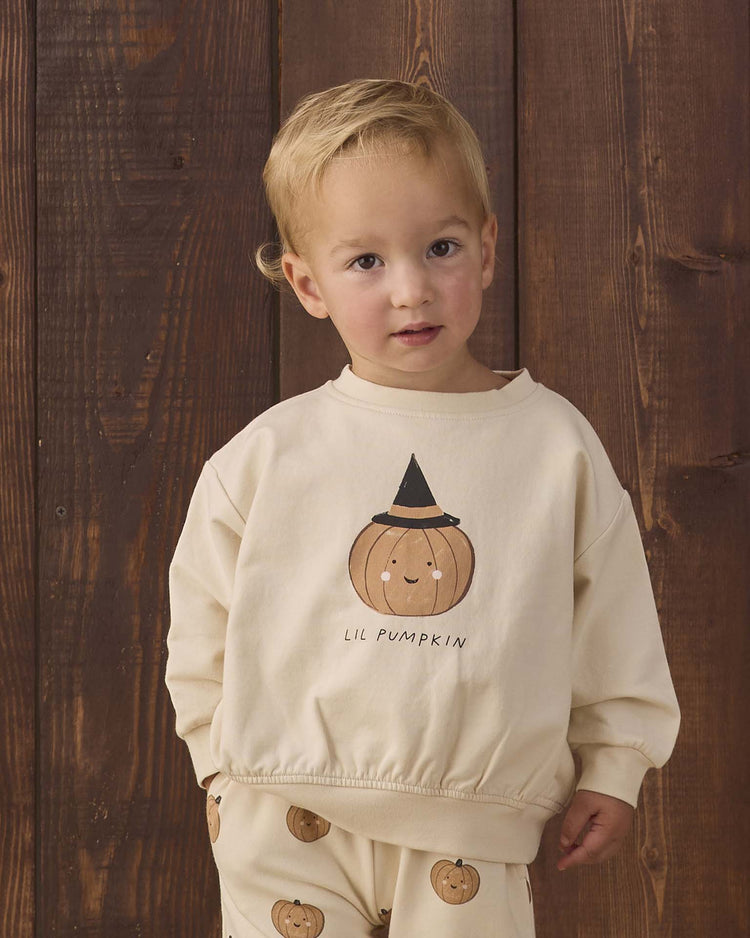 A young child stands in front of wooden boards, wearing the quincy mae relaxed fleece sweatshirt in lil pumpkin—a cozy cream organic cotton top featuring a smiling pumpkin graphic, a black hat, and the words “LIL PUMPKIN.” The look is completed with matching pants adorned with small pumpkin prints.