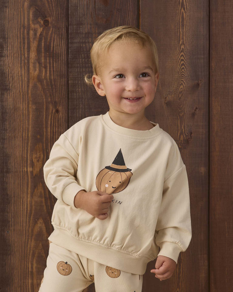 A smiling toddler wears the quincy mae relaxed fleece sweatshirt in lil pumpkin—an essential, cream-colored organic cotton top featuring playful pumpkin faces and a witch hat design—while standing in front of a wooden background.