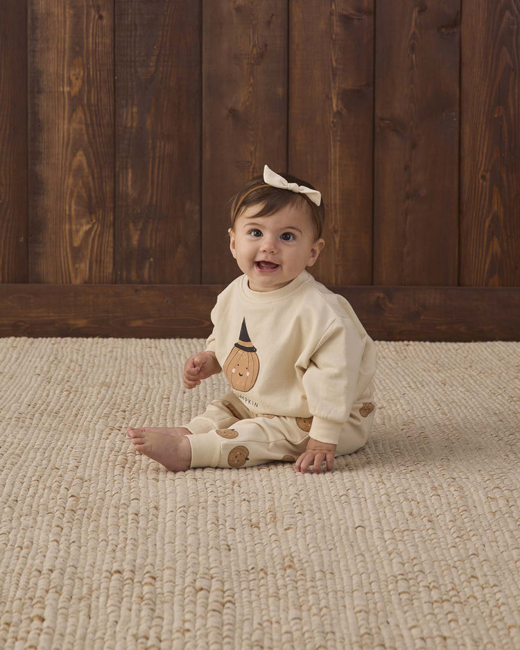 A baby wearing the Quincy Mae relaxed fleece sweatshirt in “lil pumpkin” and a matching bow sits on a textured beige rug in front of a wooden wall, smiling at the camera.