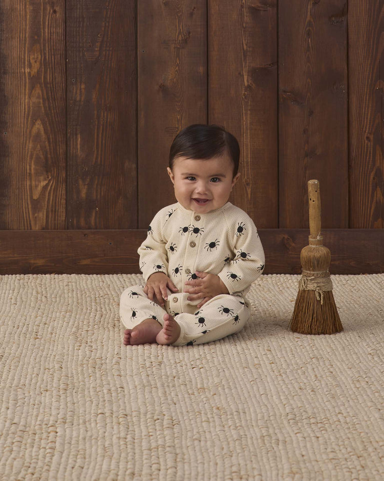 A baby wearing the quincy mae waffle long sleeve jumpsuit in bitsy spider print sits on a textured rug next to a small straw broom, with a wooden wall in the background.