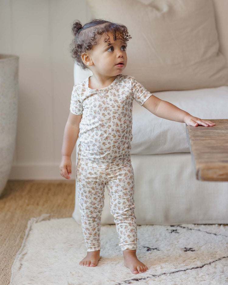 A toddler with curly hair, dressed in the quincy mae bamboo short sleeve pajama set in wildflower, stands barefoot on a soft rug and touches a wooden table while looking to the side in a cozy living room.
