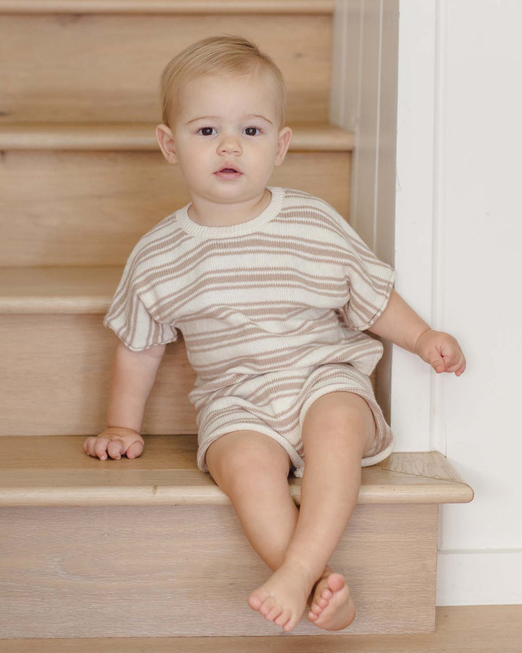 A toddler with short blonde hair sits on a light wooden staircase, wearing the quincy mae relaxed summer knit set in oat stripe. The child looks at the camera with a calm expression and has one leg stretched out on the step.