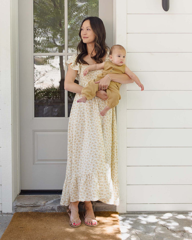 A woman in a light floral dress stands on a doormat outside a white door, holding a baby wearing the quincy mae waffle jumpsuit in honey. She gazes to the side while sunlight filters through nearby trees.