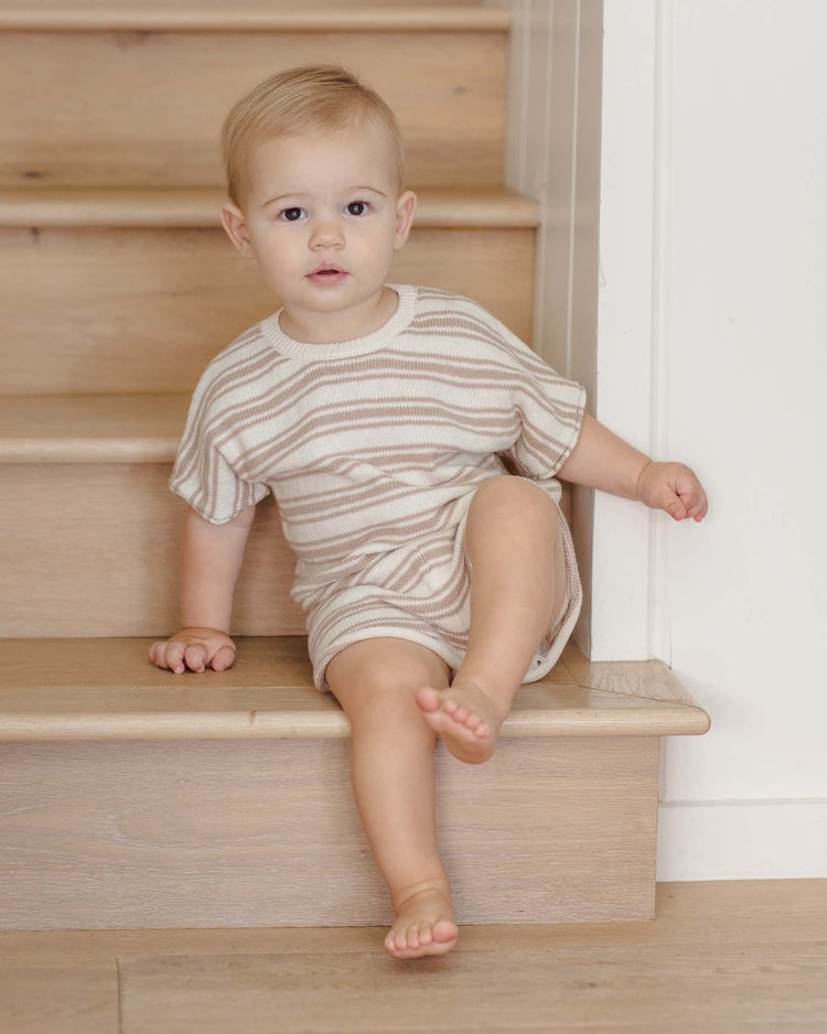 A light-haired toddler wears the quincy mae relaxed summer knit set in oat stripe—an organic cotton, beige and white striped outfit—while sitting on indoor wooden stairs with one hand on a step and one foot stretched forward.