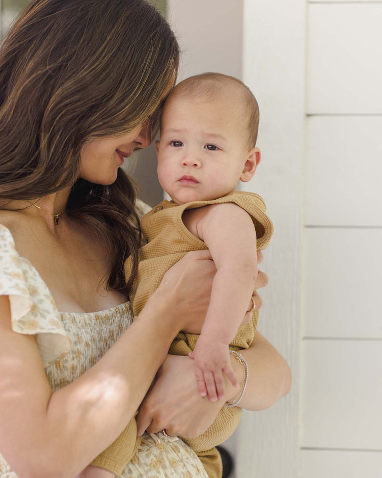 A woman in a light floral dress gently holds a baby wearing the quincy mae waffle jumpsuit in honey, standing close together in soft, natural light near a white wall.