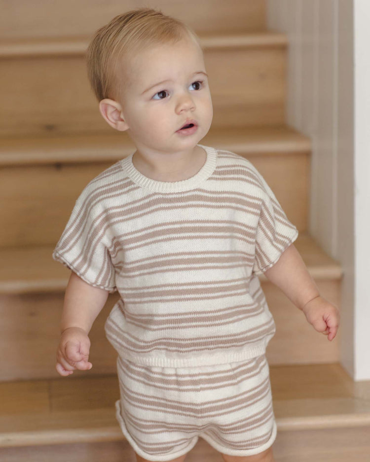 A young child with short blonde hair stands on wooden stairs, wearing the quincy mae relaxed summer knit set in oat stripe, made from organic cotton, and looks to the side with a curious expression.