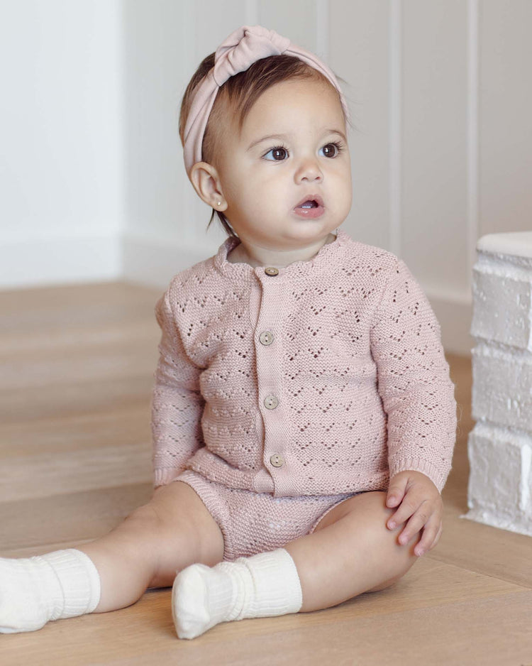 A baby girl in a quincy mae knit bloomer in blush, paired with white socks and a matching pink headband, sits on a wooden floor near a white brick wall, gazing slightly to the side.