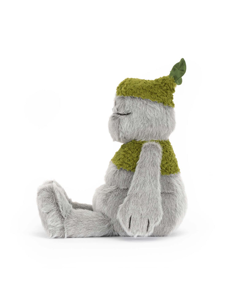 The Jellycat Rockleton is a moss plush toy with soft gray fur, wearing a green knitted hat with a leaf and a matching vest. This polyester stuffed animal sits upright in profile, eyes closed, with its arms at its sides.