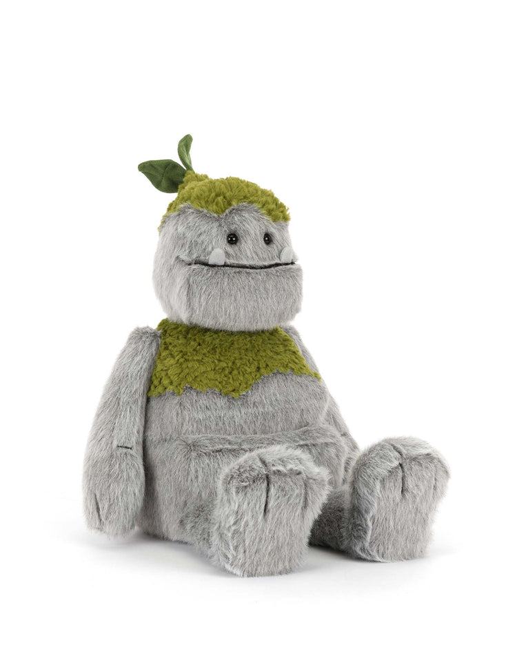 The jellycat rockleton plush features shaggy gray fur, a wide smile, small white tusks, green fuzzy patches on its head and chest, and a green leaf on top. It sits upright against a white background.