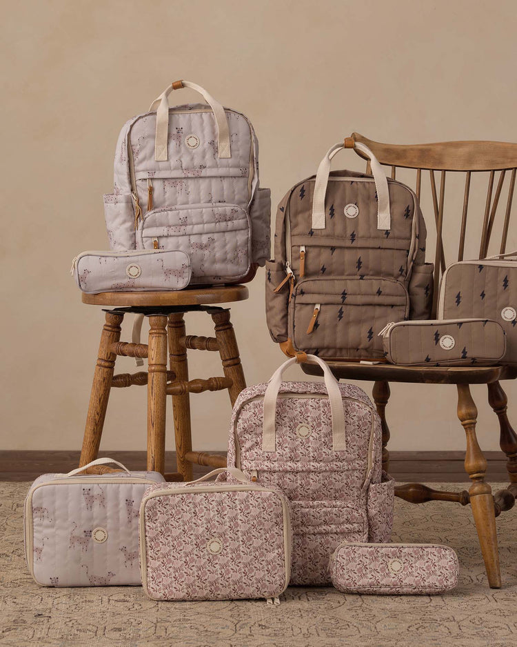 A pencil pouch in leopard by rylee + cru—crafted from durable polyester and perfect for school essentials—is displayed alongside coordinating backpacks and toiletry bags in soft beige, brown, and pink patterns on a wooden chair, two stools, and a neutral rug against a beige wall.